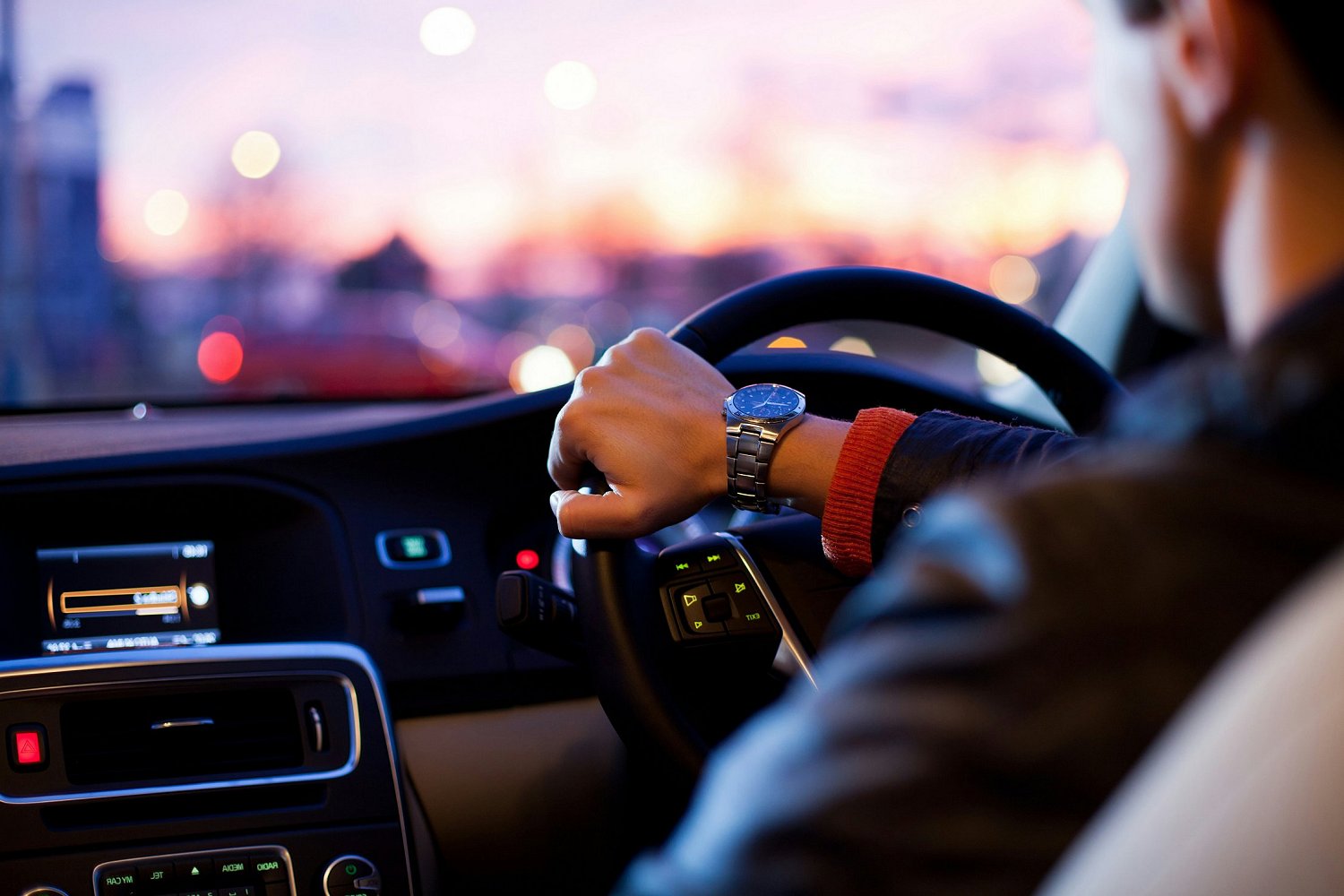 Driver steering a car at dusk with city lights in the background.