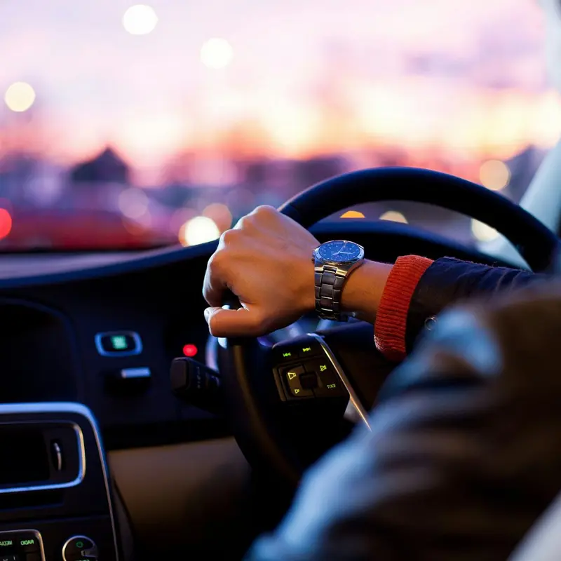 Driver steering a car at dusk with city lights in the background.