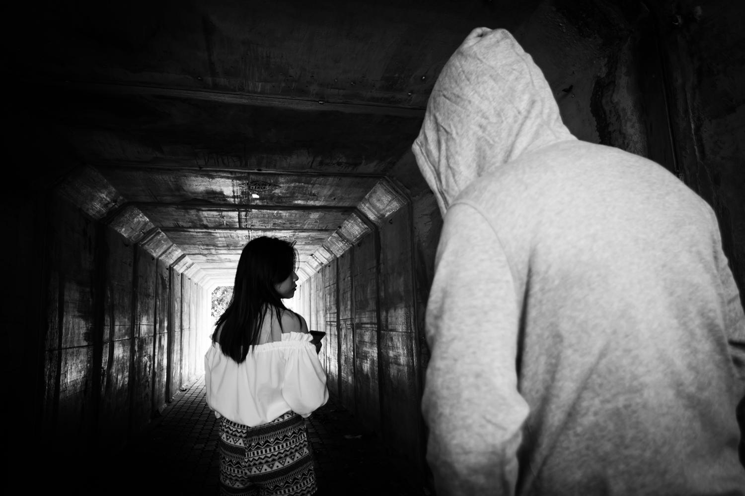 Black and white image of a hooded figure approaching a woman in a dimly lit tunnel.