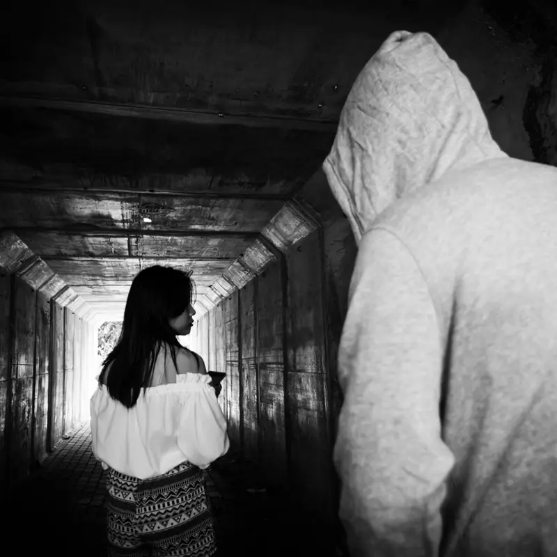Black and white image of a hooded figure approaching a woman in a dimly lit tunnel.