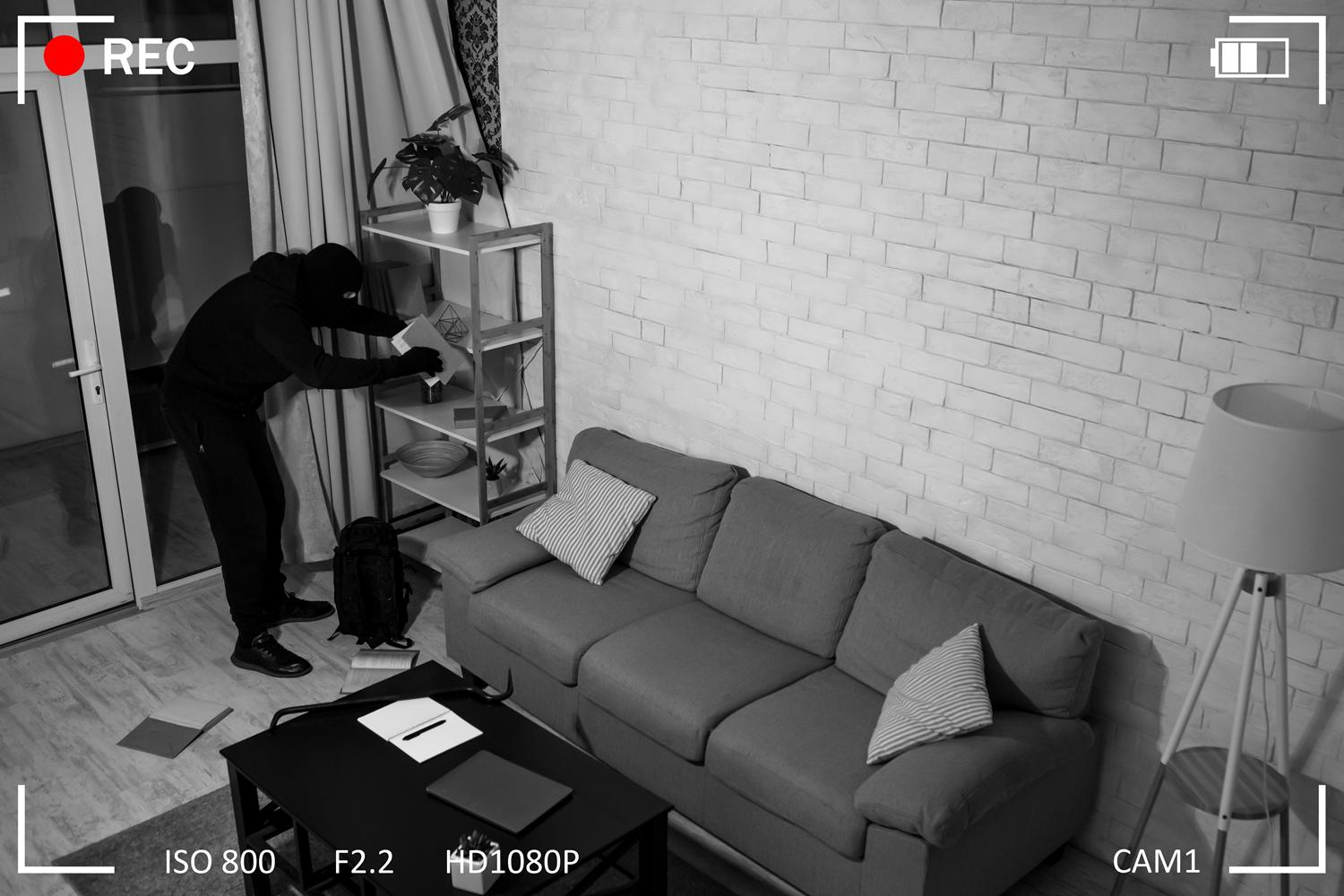 Surveillance camera footage of a masked burglar rummaging through shelves in a living room.