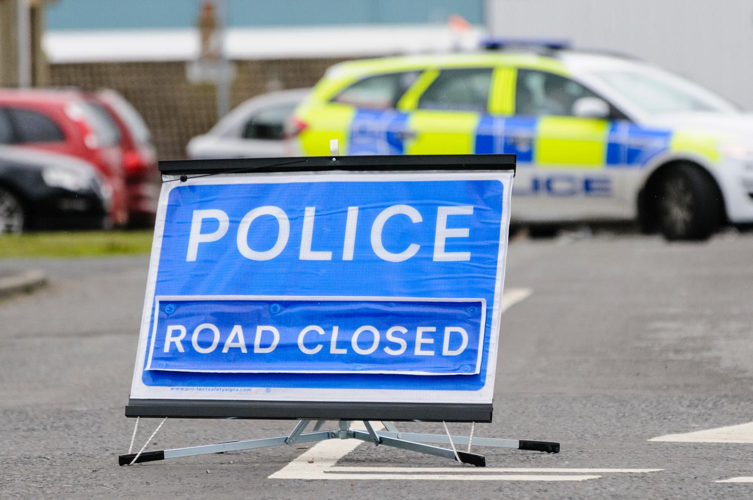 Blue 'POLICE ROAD CLOSED' sign with a police car blocking the road.