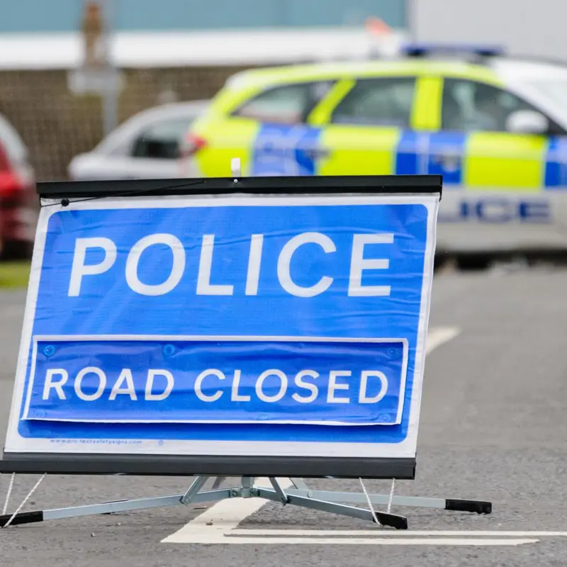 Blue 'POLICE ROAD CLOSED' sign with a police car blocking the road.