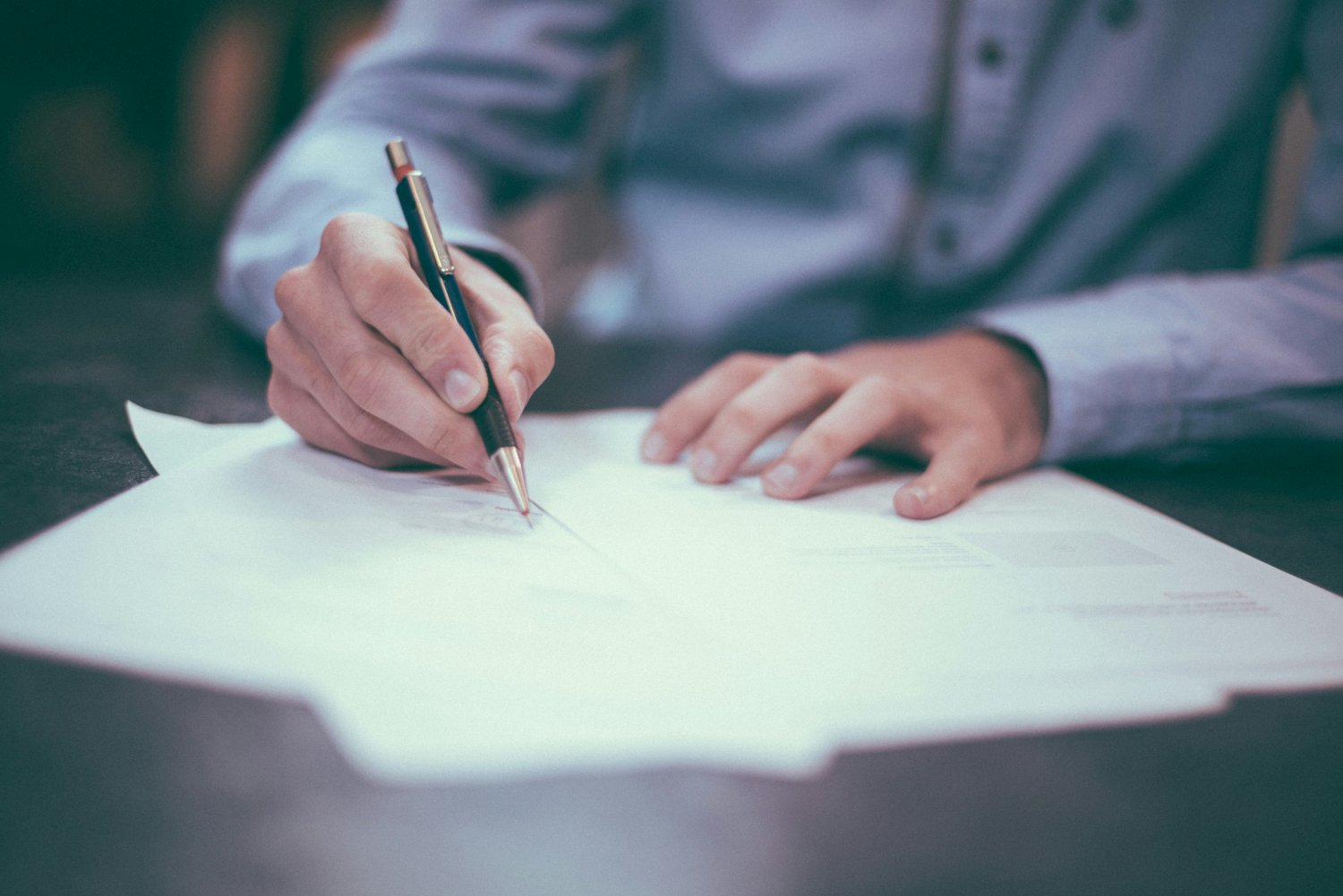 Person in a dress shirt signing documents with a pen on a desk.