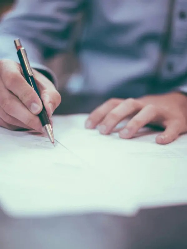Person in a dress shirt signing documents with a pen on a desk.