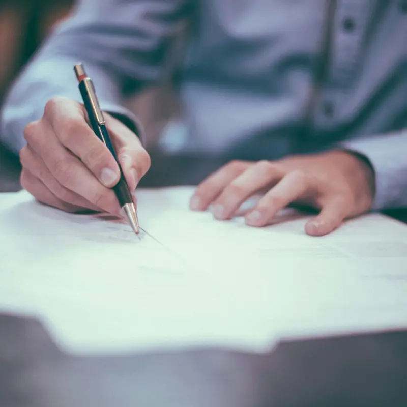 Person in a dress shirt signing documents with a pen on a desk.