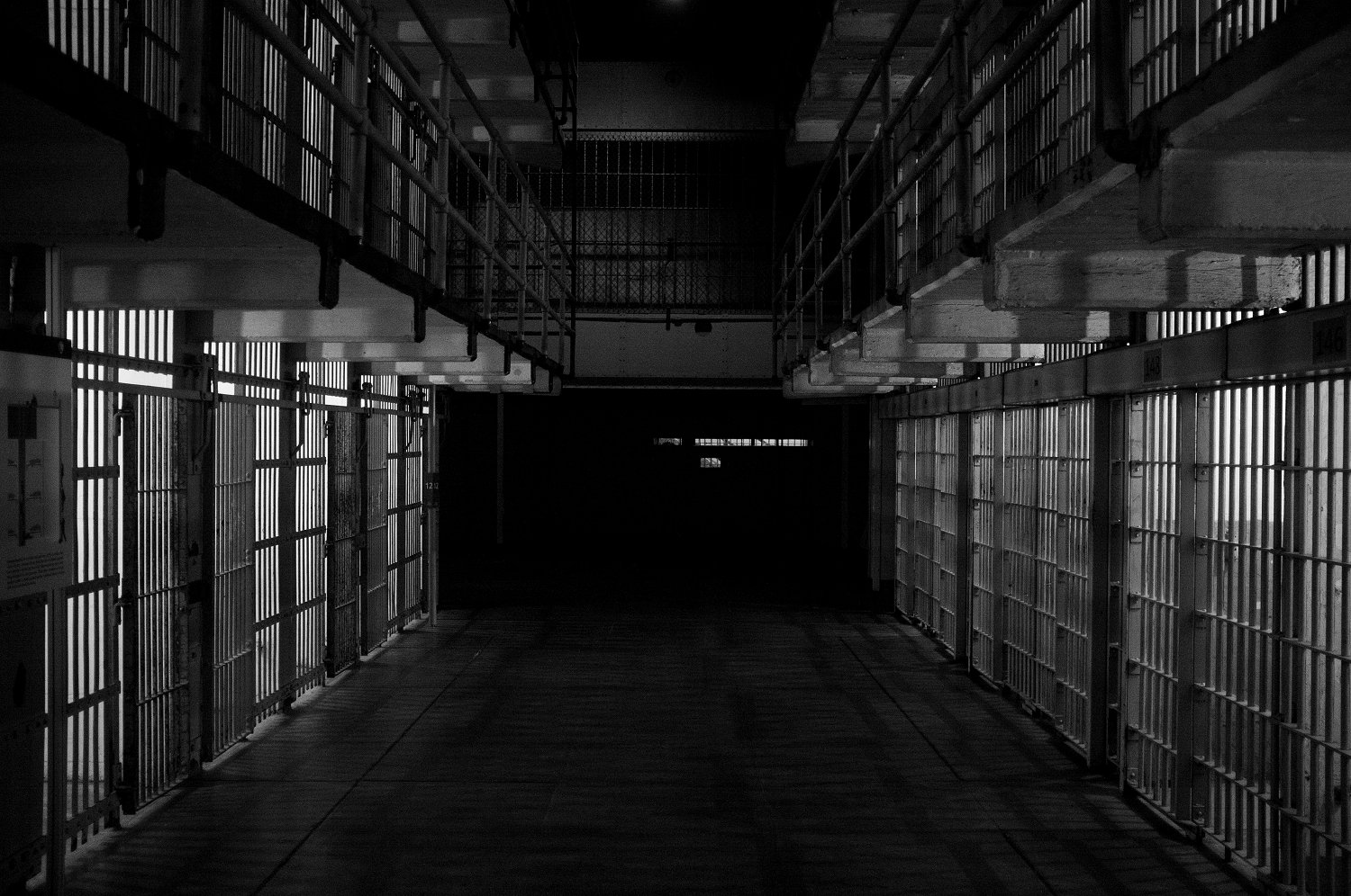 Dimly lit prison corridor with rows of metal-barred cells on both sides.