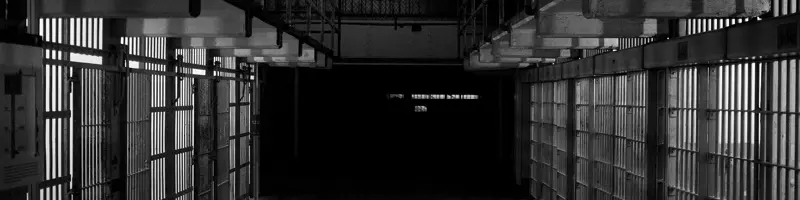 Dimly lit prison corridor with rows of metal-barred cells on both sides.