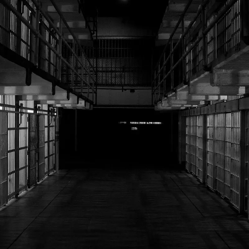 Dimly lit prison corridor with rows of metal-barred cells on both sides.