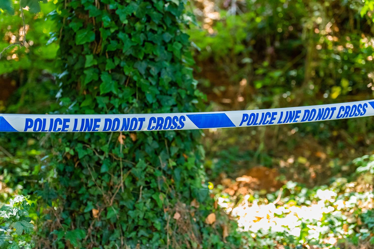Blue 'POLICE LINE DO NOT CROSS' tape cordoning off a wooded area.