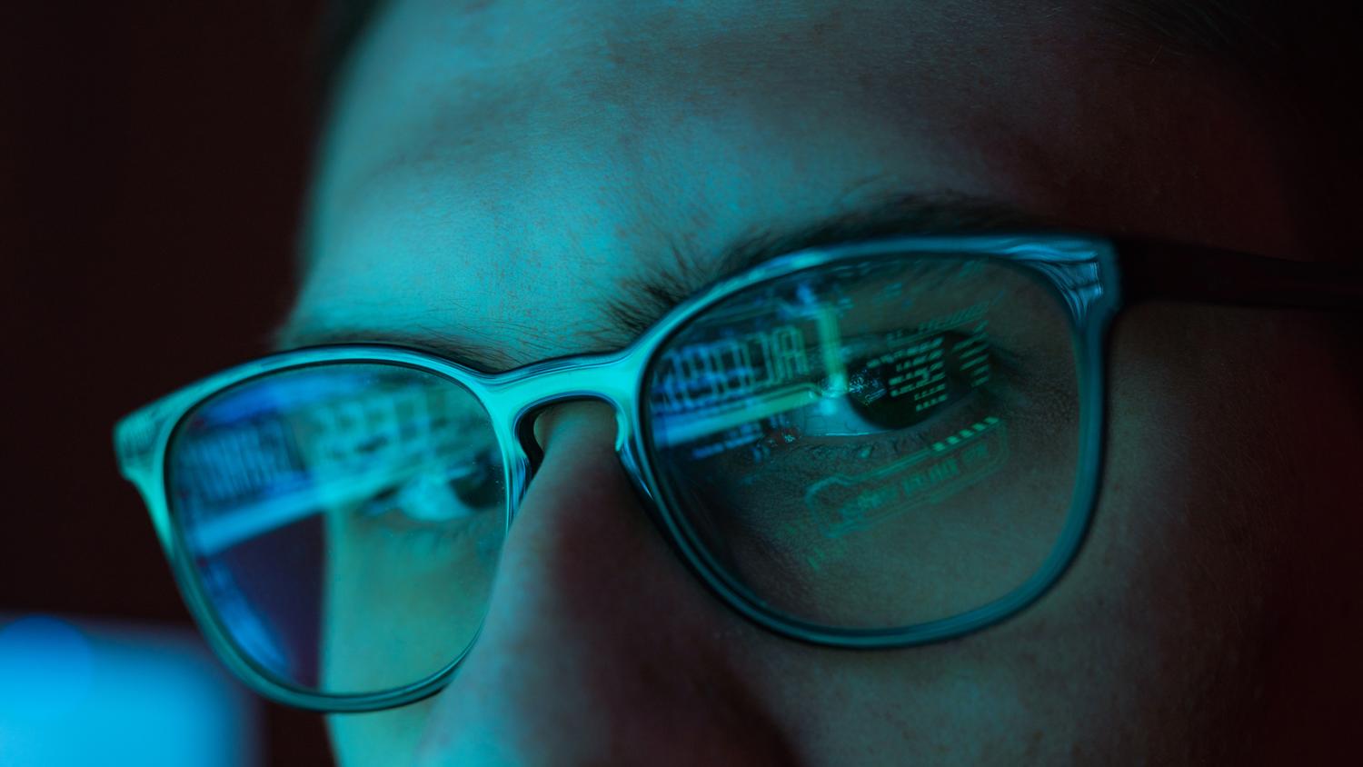 Close-up of a person wearing glasses with computer code reflecting in the lenses.