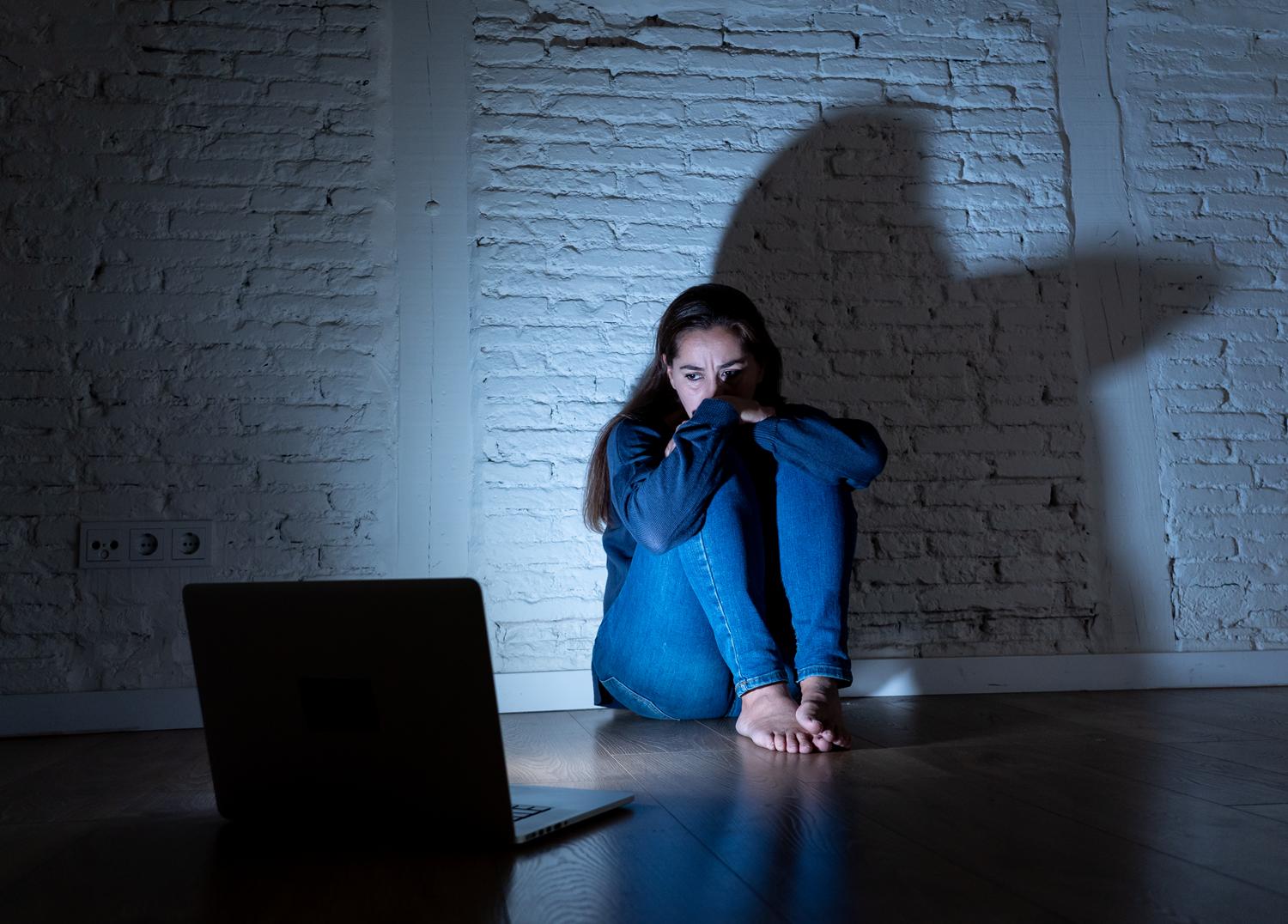 Young woman sitting in fear near a laptop, illuminated by a shadowy figure on the wall.