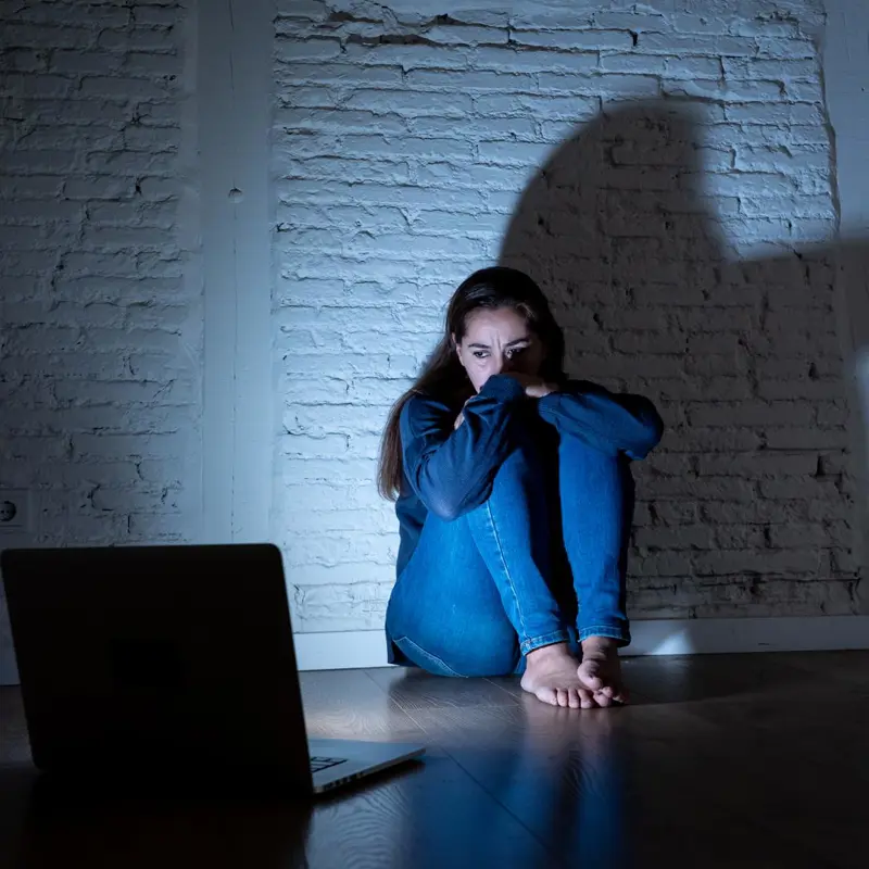 Young woman sitting in fear near a laptop, illuminated by a shadowy figure on the wall.